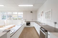 the nursery kitchen is very modern and bright with white units and dark wood worktops. there are fivesinks, an oven and a dishwasher. the serving area is low down and beside a long dining table