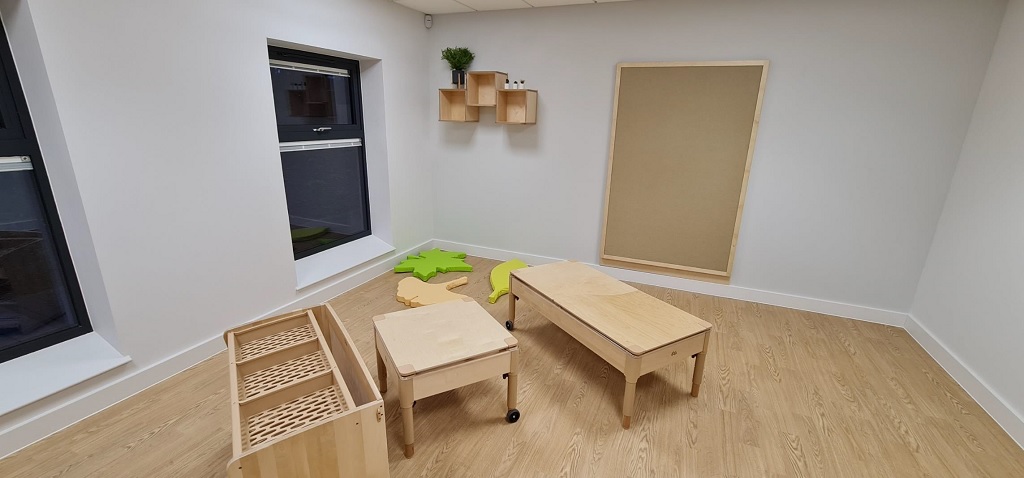 A room with natural wood play furniture, a pinboard on the wall and some box shelves
